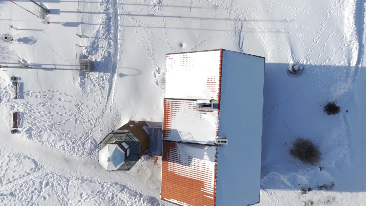 A crisp bird's-eye view of a winter settlement featuring buildings with red roofs and a small tower. Footprints and vehicle tracks mark the pristine white snow around the structures