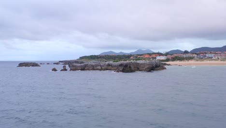 Drone-flyover-Atlantic-Ocean-toward-small-rocky-islet-packed-with-birds,-waterfront-town-in-background