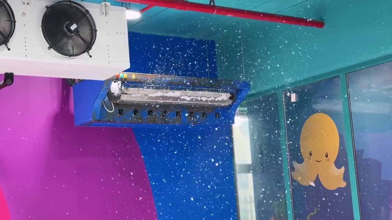 A blue artificial snow machine suspended from the ceiling drops white foam flakes inside a brightly colored indoor children's play area featuring a cute octopus mural.