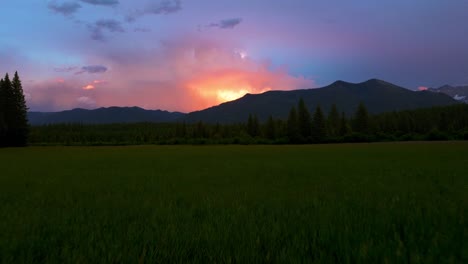 épica-Puesta-De-Sol-De-Verano-Sobre-El-Paisaje-Montañoso-De-Montana-Durante-La-Tormenta-De-Lluvia-Con-Relámpagos-Destellando-A-Través-De-Las-Nubes-Mientras-La-Cámara-Empuja-Sobre-Un-Exuberante-Campo-Verde-De-Hierba-Bordeado-De-árboles-Altos