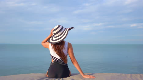 A-woman-with-her-back-to-the-camera,-dressed-in-a-black-and-white-bathing-suit-and-matching-sun-hat,-sits-on-a-cushioned-seat-looking-out-at-the-ocean-horizon