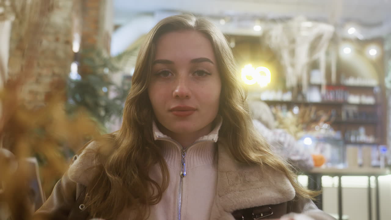 Portrait shot of young woman smiling as she rests face on hands in cozy cafe, peaceful moment captured with soft lighting, creating serene and warm atmosphere in relaxing environment