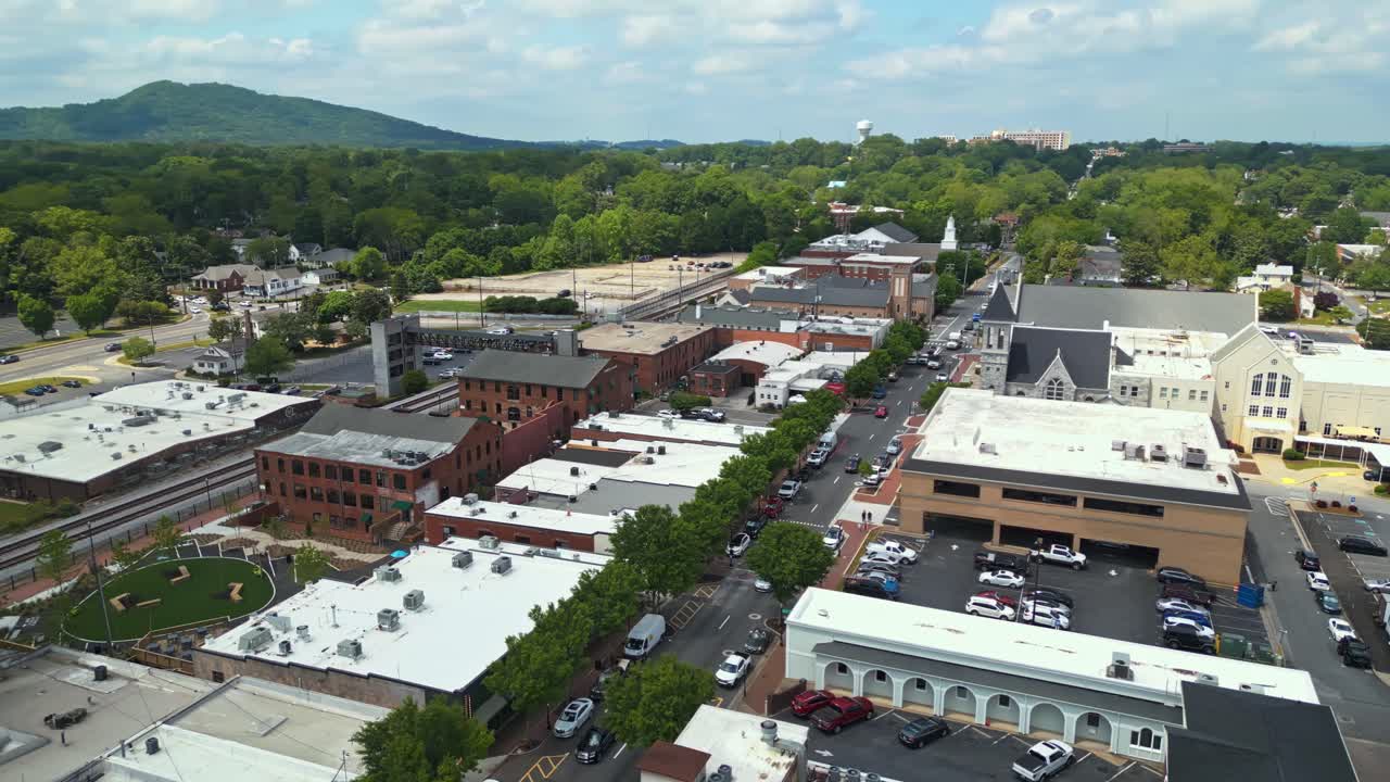 Aerial wide shot of main street in suburbia of Marietta City with green trees and mountains in distance. Tree-lined avenue with stores and shops in spring.