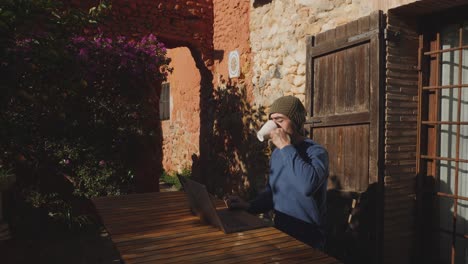 a man working outside a cottage working with his laptop, makes a pause and drinks coffee