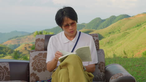 portrait of asiatic male reading a book sitting outdoor in leather coach vintage sofa with mountains scenic view on the background
