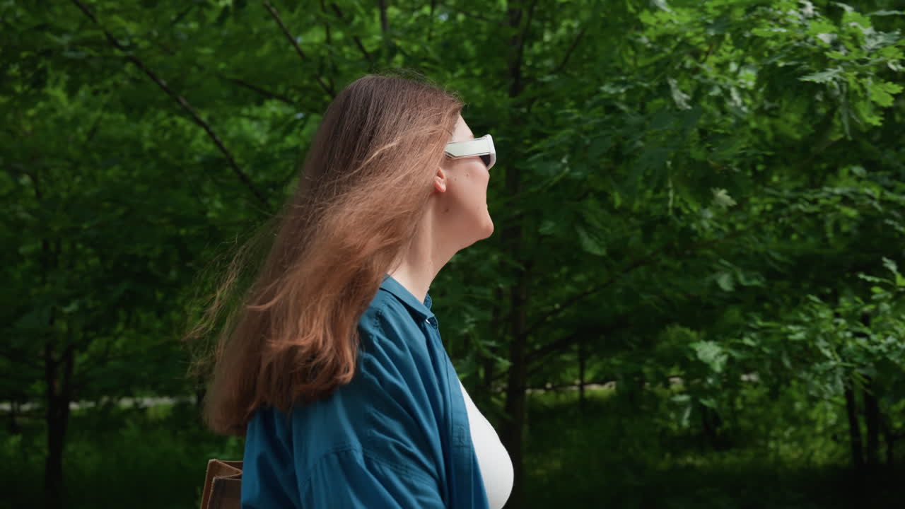 여름 햇빛 아래 선글라스와 파란색 셔츠를 입고 울창한 녹색 잎으로 둘러싸인 숲길을 따라 한가로이 산책하며 평화로운 야외 산책을 즐기는 관광객의 측면 모습