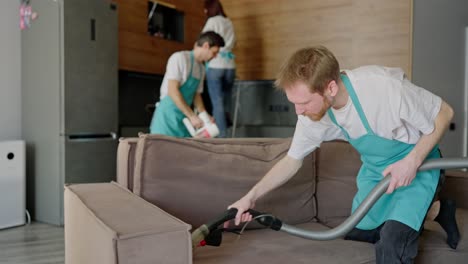 Side-view-of-a-confident-trio-group-of-cleaners-in-white-T-shirts-and-blue-aprons-wiping-down-an-apartment-and-a-man-using-a-vacuum-cleaner-cleaning-a-gray-sofa-in-a-modern-apartment-cleaning-on-call