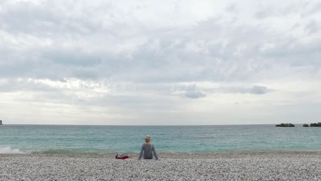 woman sitting on a pebble beach, looking at the ocean