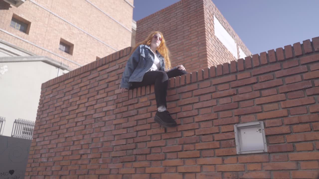 Two friends have fun taking pictures in the street. Redheaded model poses sitting on top of a brick wall and smiles for her photographer friend