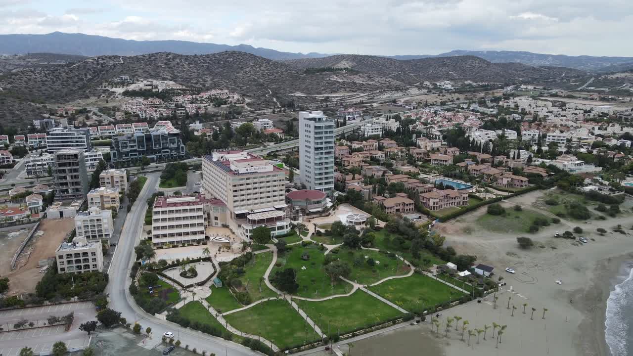 Drone shot of green beachfront park next to tall high-rise building, with winding shoreline and residential area extending toward distant hills under cloudy sky