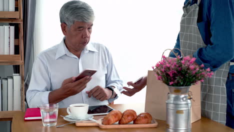 Active-senior-Asian-man-working-in-a-coffeeshop-using-a-mobile-device,-smartwatch-for-NFC,-QR-code-scanning,-contactless-payment-to-pay-for-coffee-and-snack