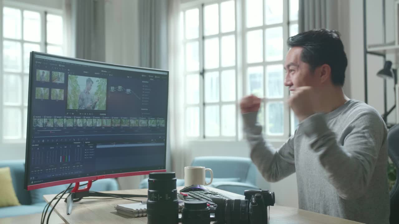 editor de video asiático hombre con camiseta de mangas largas siendo feliz terminando la edición de video por computadora de escritorio mientras trabaja en casa.