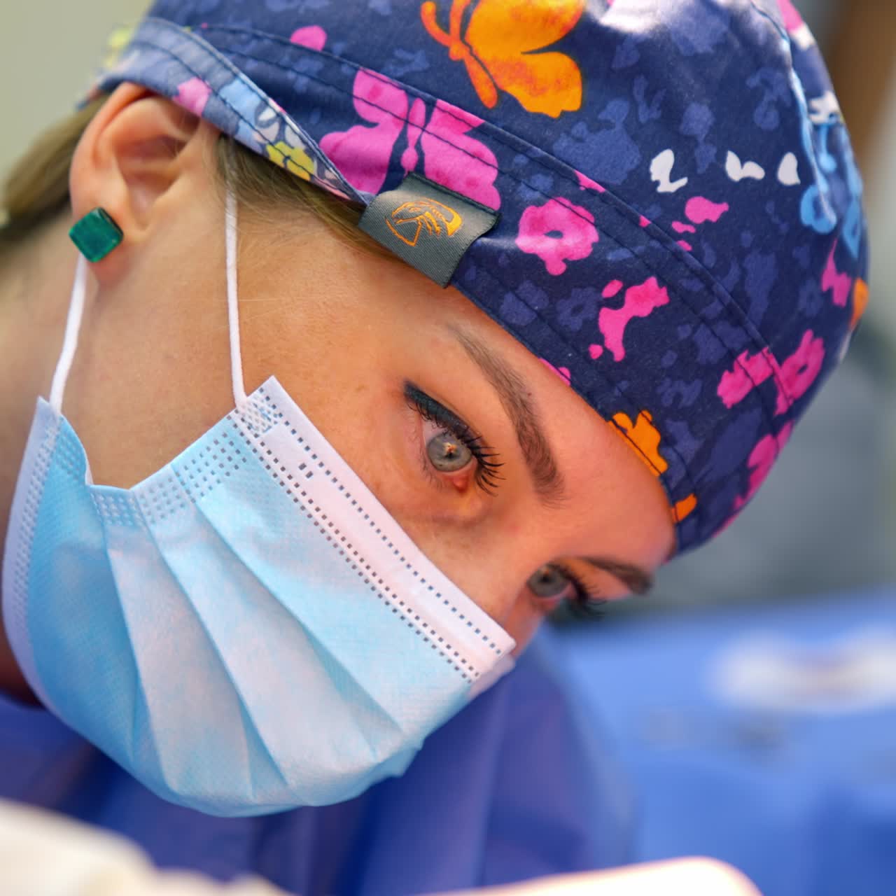 Female doctor in mask and colorful hat during operation. Woman surgeon looks intently on the operated area. Close up portrait