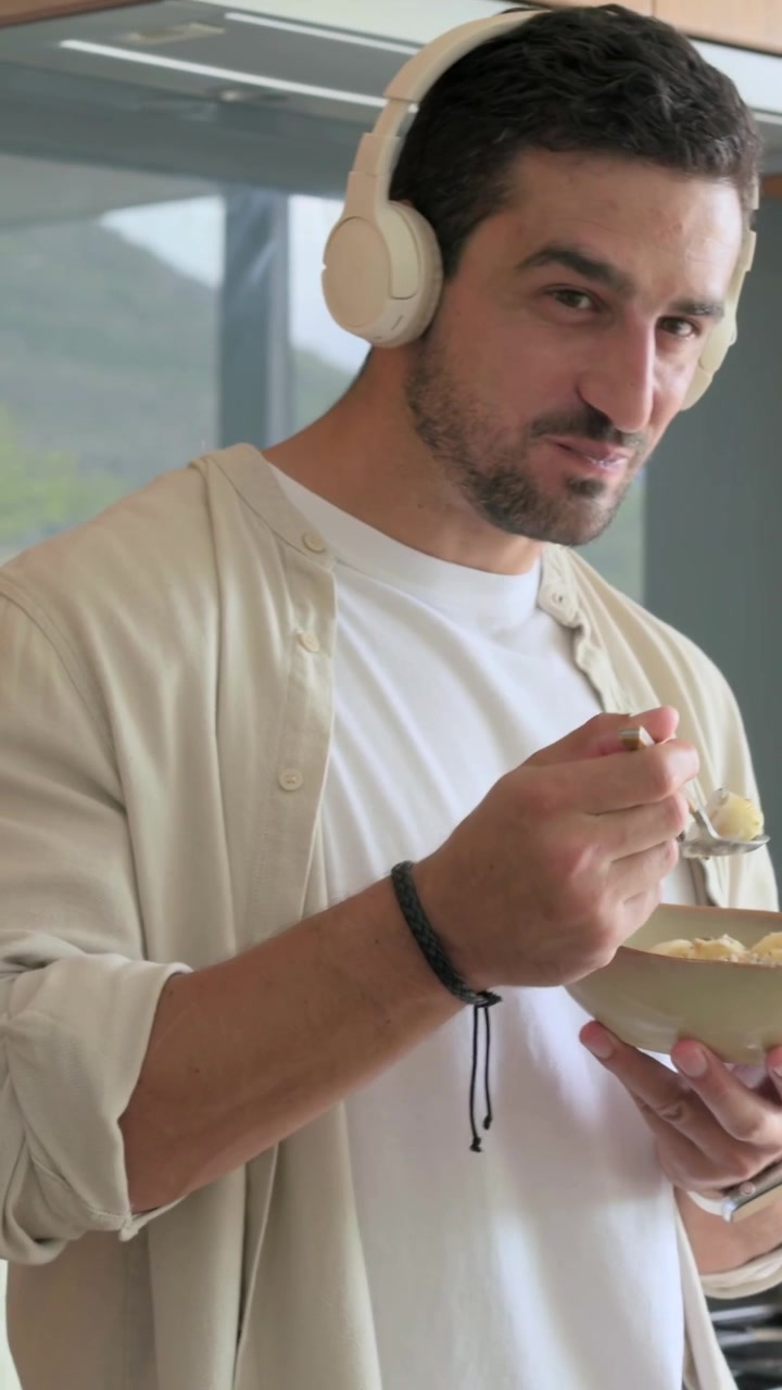 Vertical video: Listening with headphones, man sampling with spoon at kitchen window, holding bowl