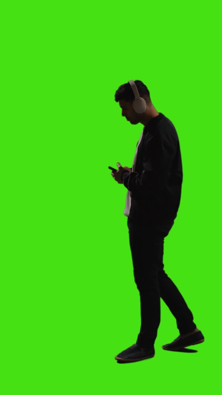 Video verticale: ripresa a figura intera di un uomo con cuffie wireless che ascolta musica in streaming dal cellulare, su sfondo green screen 1