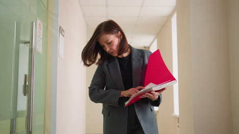 businesswoman reviewing documents in red folder, walking down office hallway, corporate environment, professional attire, focused on work, reading papers, modern workspace, office walk