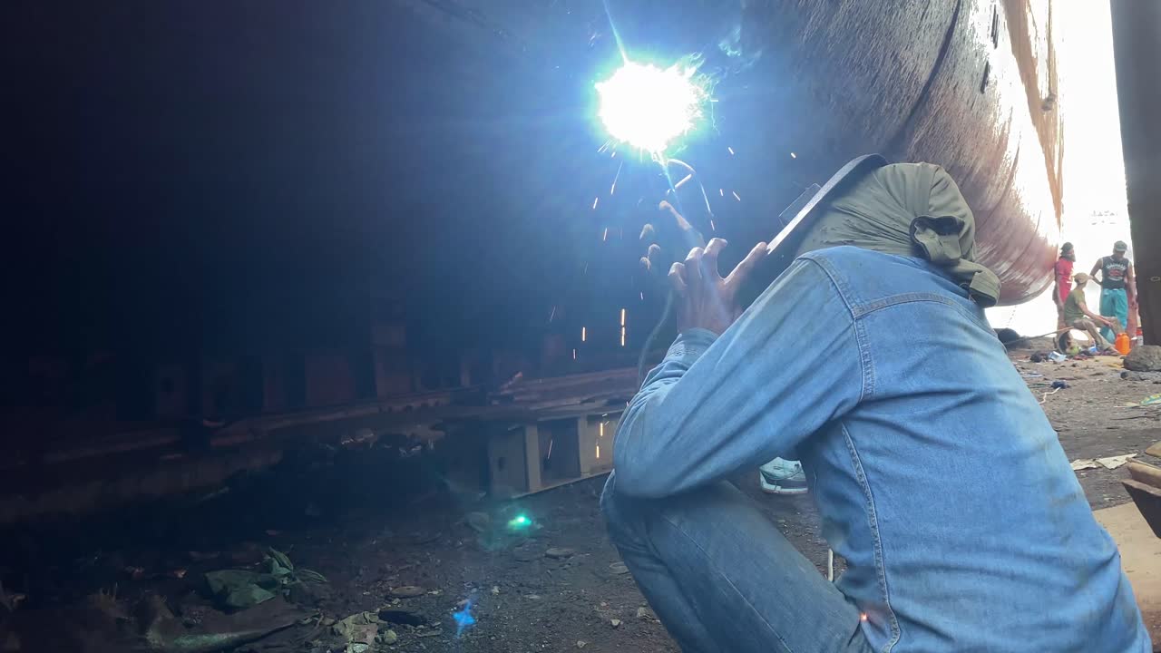 Close up shot of a welder worker man welding hull of a black ship in shipyard at daytime