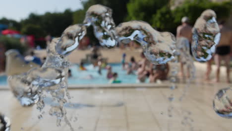 Fuente-De-Agua-En-Cámara-Lenta-En-Un-Camping,-Gente-Relajándose-En-El-Fondo-De-Una-Piscina