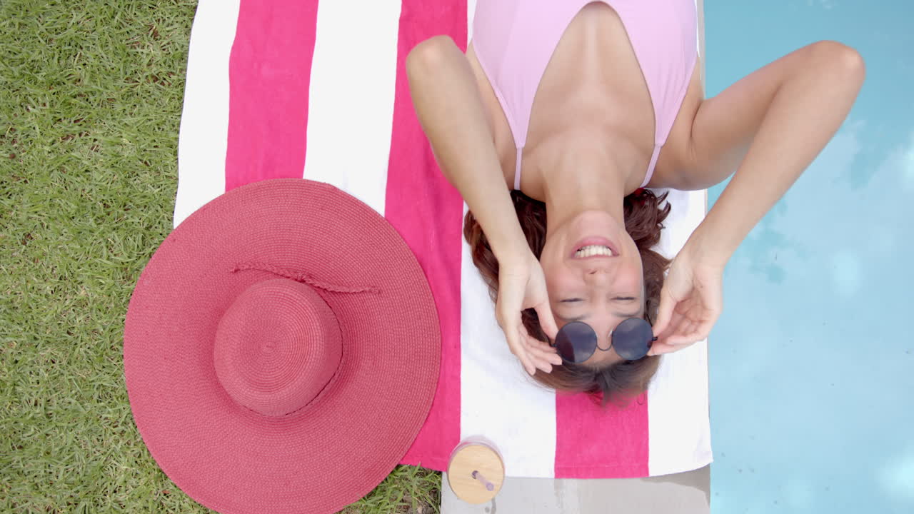 Young biracial woman in a pink swimsuit relaxes on a striped towel by the pool