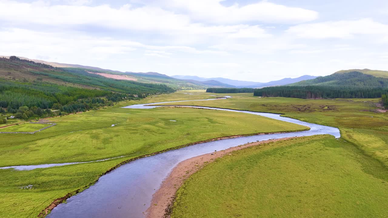 Drone camera glides above winding river, lush green grassland, and hills under soft daylight