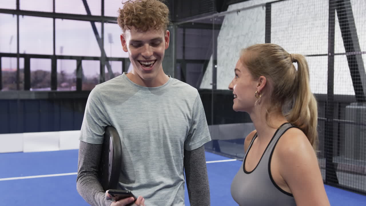 Young man and woman smiling, checking smartphone after padel tennis match on indoor court
