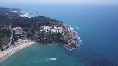 Luftaufnahme-Von-Booten,-Die-Auf-Dem-Arabischen-Meer-Um-Das-Leela-Raviz-Kovalam-Hotel-In-Kerala,-Indien-Herumfahren-–-Drohnenrückzug