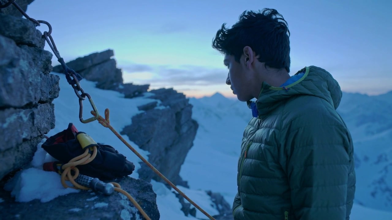 Jovem escalador vestindo jaqueta verde inspecionando cordas e ancoragem em uma borda nevada, equipamento