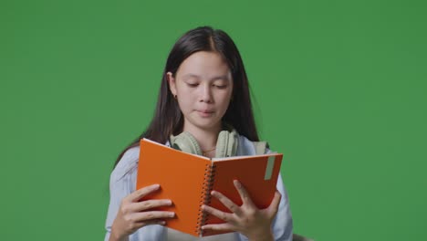 close up of asian teen girl student with a backpack holding and reading a book while walking in the green screen background studio