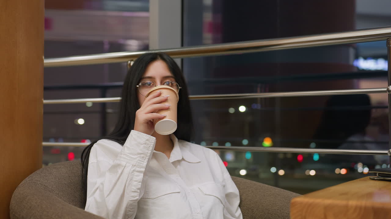 young woman with glasses seated on chair sips hot drink with satisfaction while enjoying calm moment in modern interior, with night city lights visible through glass panel in background