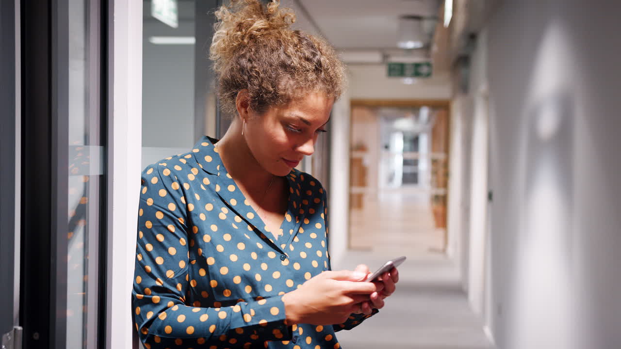 Mixed race millennial woman leaning against a wall in an office corridor using smartphone, selective focus