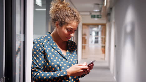 Mixed-race-millennial-woman-leaning-against-a-wall-in-an-office-corridor-using-smartphone,-selective-focus