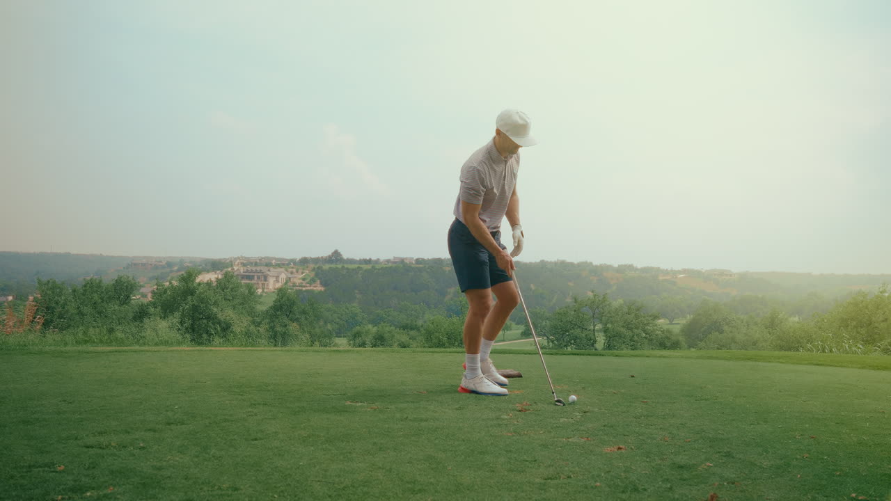 A male golfer walks up to the ball on the tee at a scenic hilltop course. The natural landscape and elevated view create a peaceful, cinematic moment of focus before the swing.