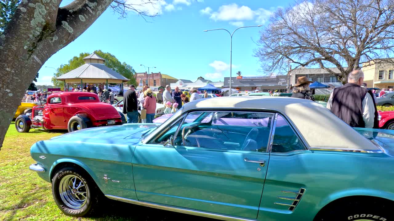 Vintage cars, including a turquoise coupe, are showcased on a sunny day at a lively outdoor festival with crowds and historic buildings in Tregony, Queensland