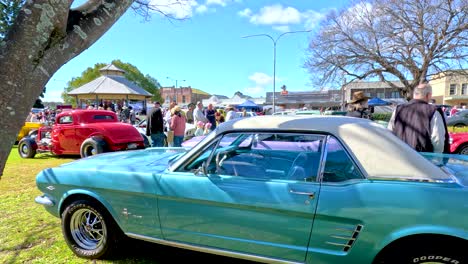 vintage cars, including a turquoise coupe, are showcased on a sunny day at a lively outdoor festival with crowds and historic buildings in tregony, queensland