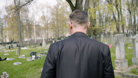 vista trasera de un hombre con traje negro sosteniendo rosas rojas y caminando en un cementerio