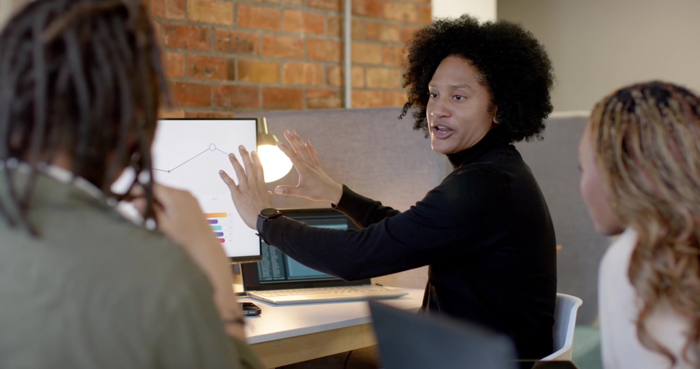 African American desk team watching woman in black top pointing at monitor trend and seeking input
