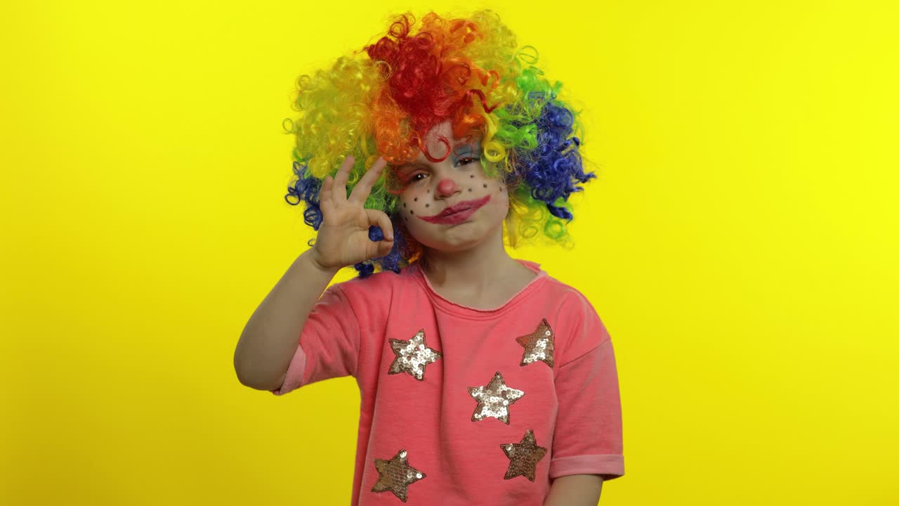 una niña payasa con una peluca colorida haciendo caras tontas. muestra el signo ok, sonriendo. halloween