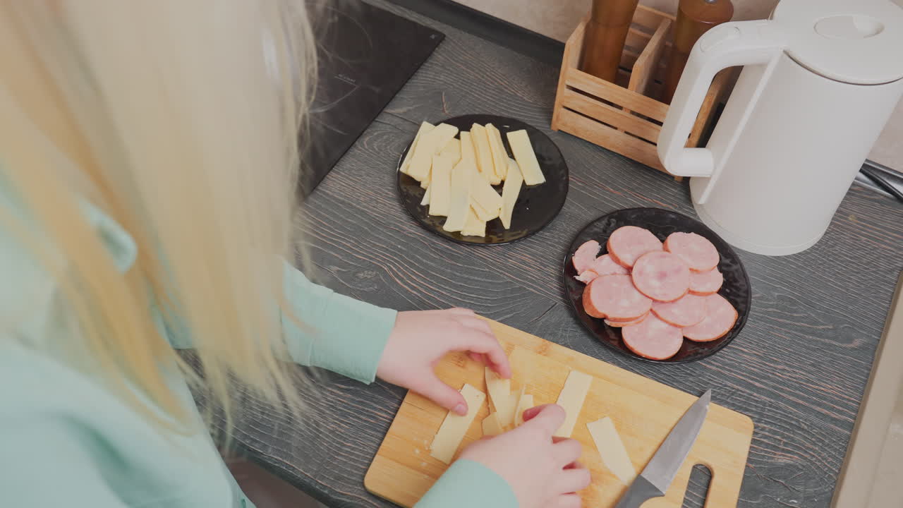 overhead view of blonde woman wearing green shirt placing sliced yellow cheese onto black plate beside sliced sausage while preparing food in kitchen with kettle, knife, and spice holder