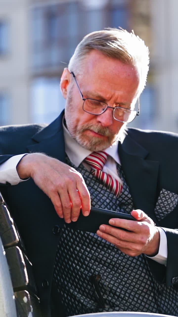 Mature businessman with beard and glasses sitting on bench in the city. Middle aged man looking seriously into his mobile phone outdoors. Vertical video