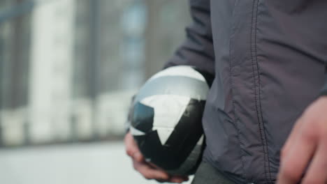 close-up side view of someone walking in sports environment holding black and white soccer ball, showing athletic wear and part of body, with urban background