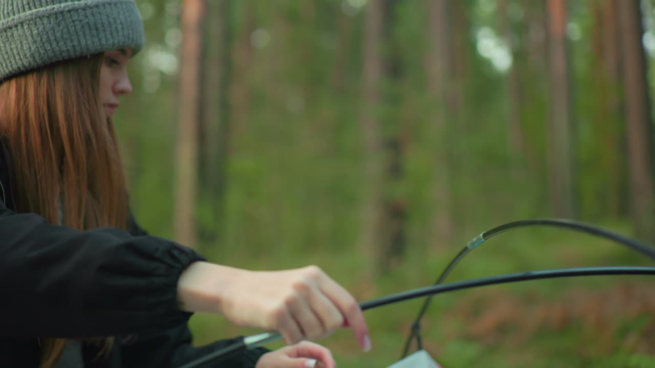 man and woman work together to assemble camping tent in lush forest as one lifts flexible pole and other secures fabric on grassy ground surrounded by tall trees and calm outdoor atmosphere