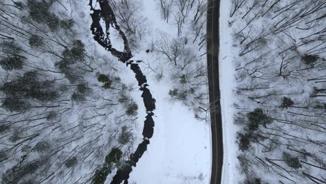 Birds-eye-view-of-a-snowy,-remote-mountain-road-with-a-stream-and-forest