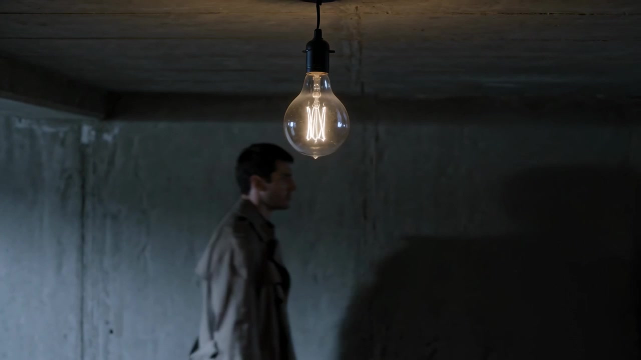 Entering left, man in trench coat walking across concrete room casting shadow under bulb to doorway