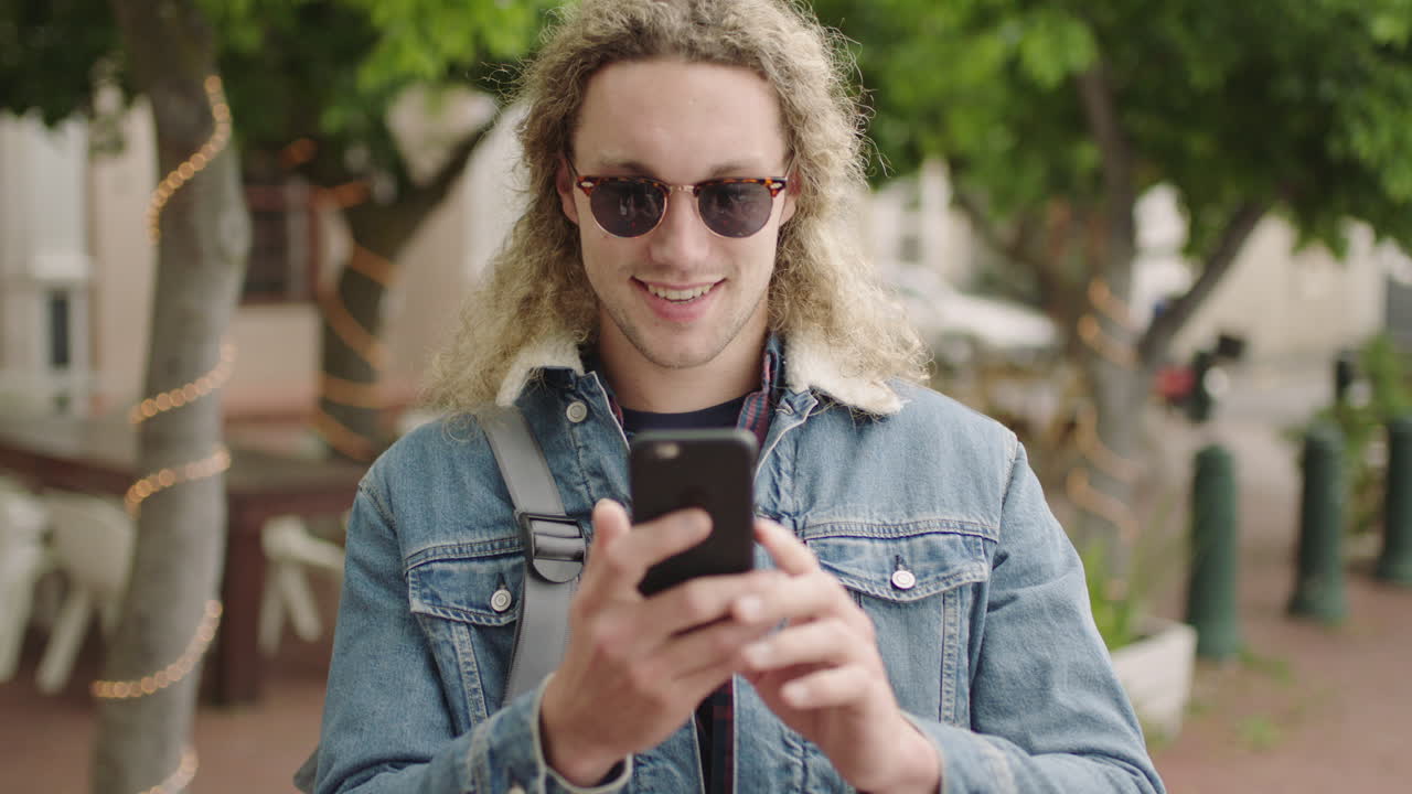 ritratto di un attraente giovane studente maschio biondo che indossa occhiali da sole, invia messaggi, naviga con lo smartphone, ride felice, gode della tecnologia mobile in città