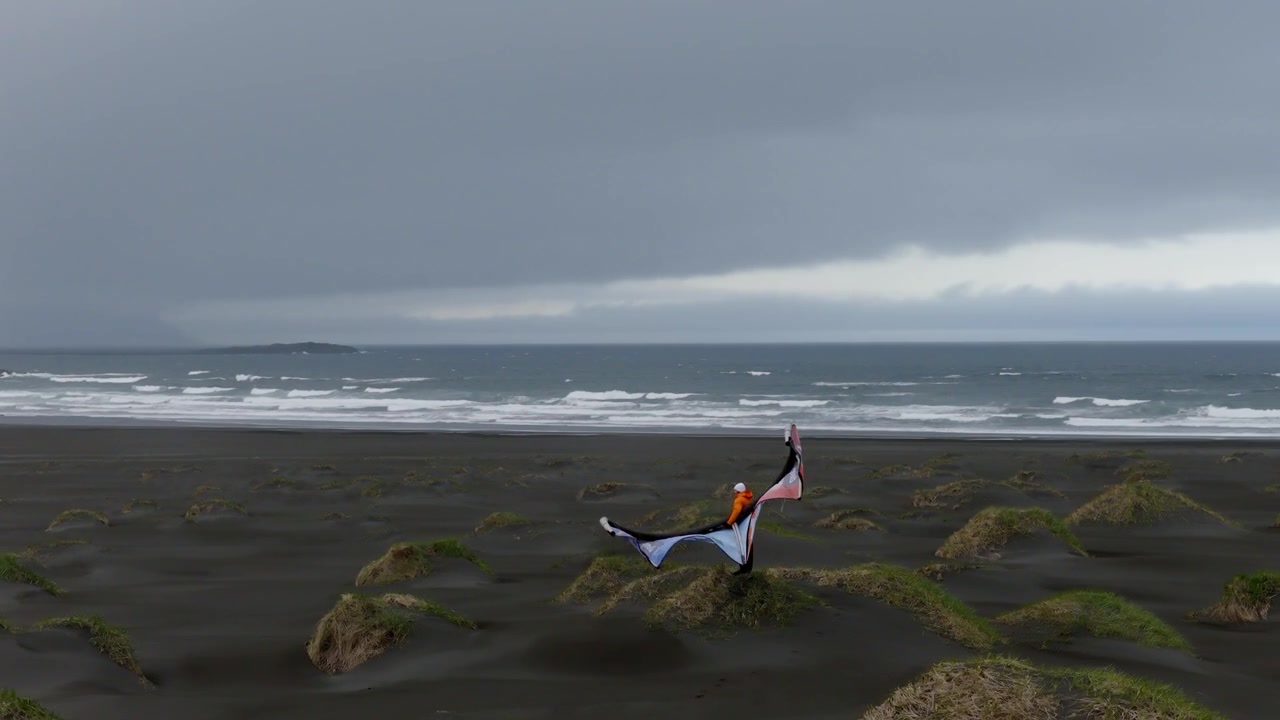 Kite surfer handles inflated kite and equipment across moss covered black volcanic sand, with strong coastal wind and Atlantic surf at Stokksnes, Southeast Iceland, drone 360 orbit.