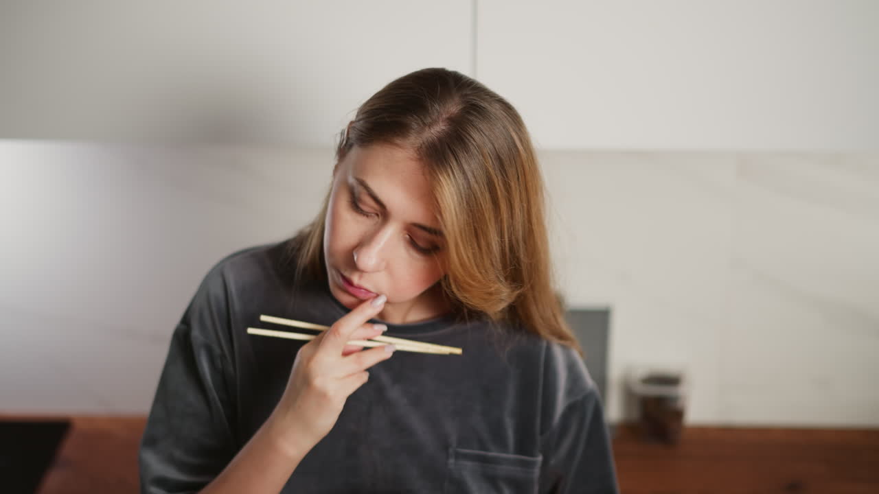 Portrait view of young housewife in gray outfit eating breakfast noodles with chopsticks, focused as she removes something from mouth, standing in bright kitchen with slightly blurred background