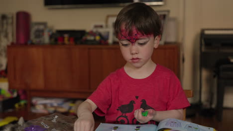 Static-shot-of-a-boy-with-red-face-paint-building-a-House-Out-of-Colored-Blocks-of-Lego-at-the-Table