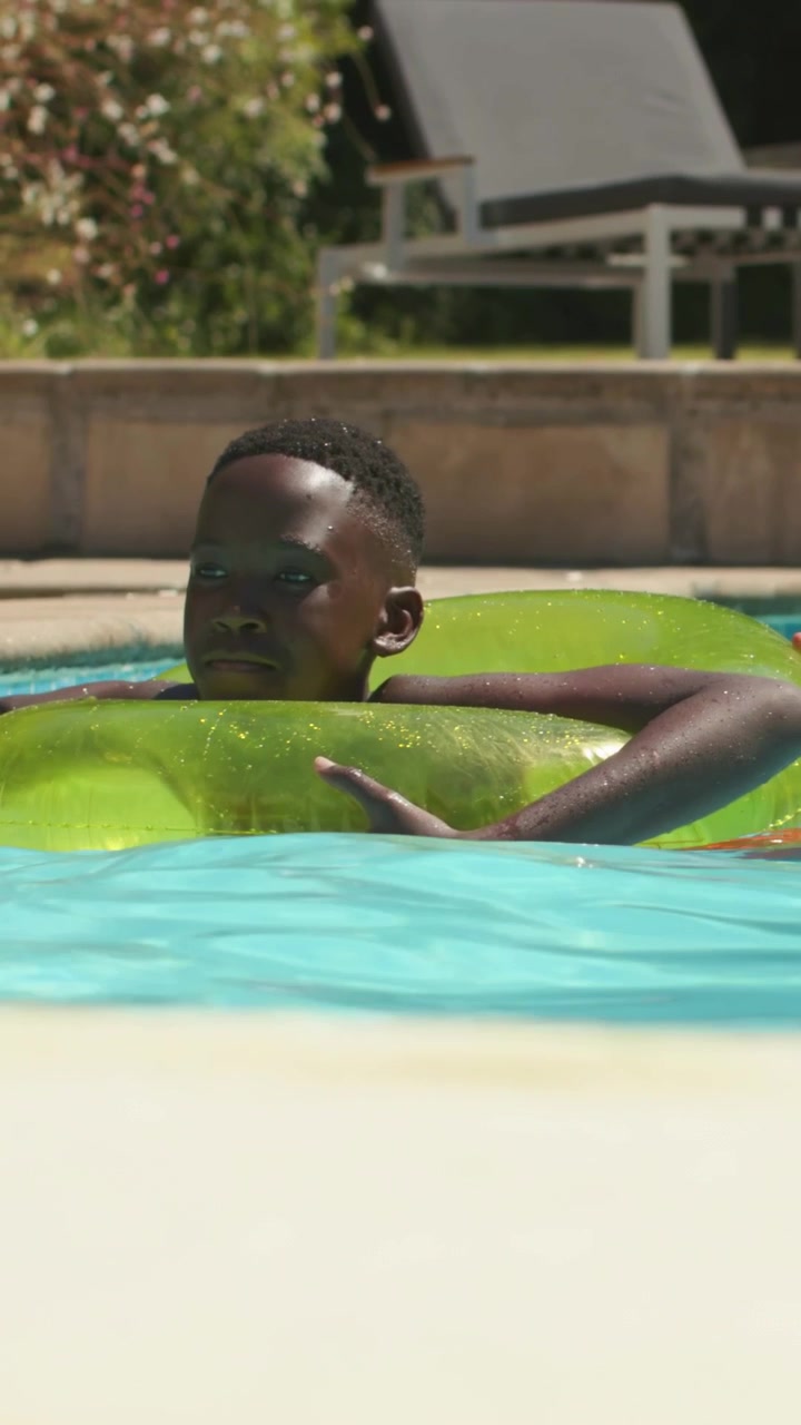Vertical video: Starting African youth in trunks at home pool edge, paddling lime tube, relaxing