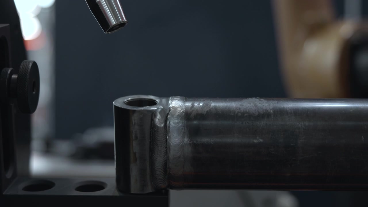 Torcia di saldatura lucida aleggia sopra un tubo d'acciaio con saldatura e hardware di supporto all'interno di un'officina buia, mostrando una texture metallica riflettente, toni freddi, profondità di campo ridotta, vista laterale ravvicinata e precisione.