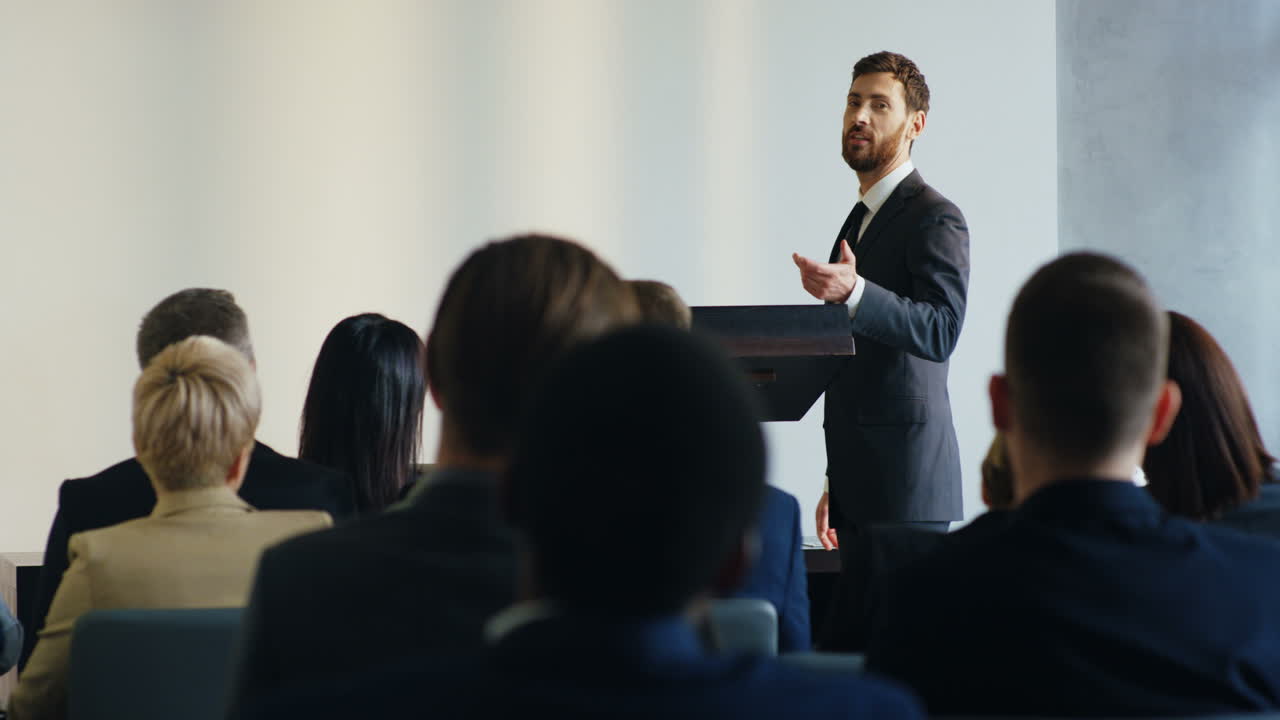 Caucasian businessman speaker on a podium wearing formal clothes and talking in a conference room in front to many people